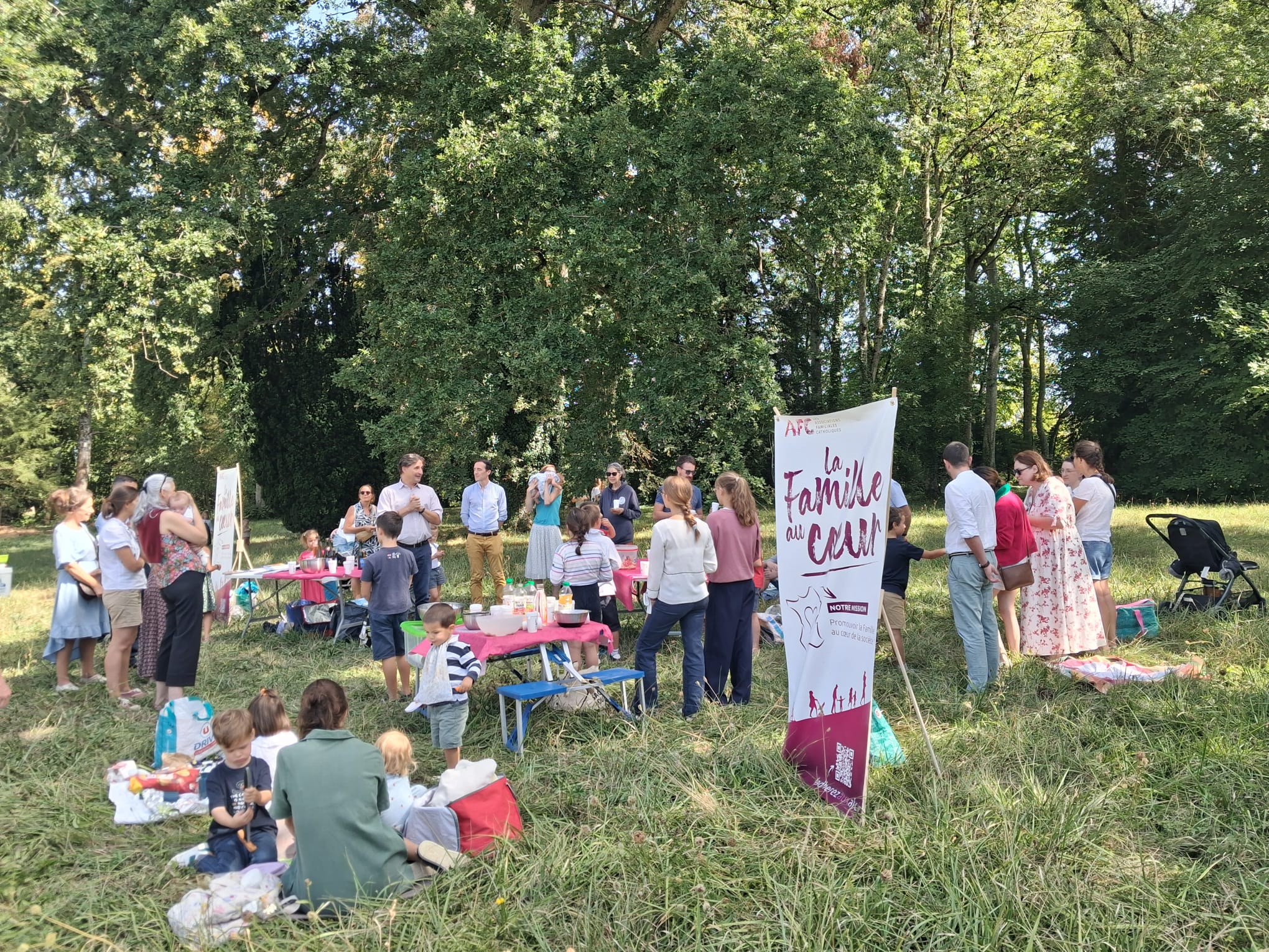 Familles de l’AFC du Pays de Rennes réunies autour des tables du pique-nique de rentrée, sous les arbres du domaine de Blossac, avec le kakémono « La Famille au cœur »